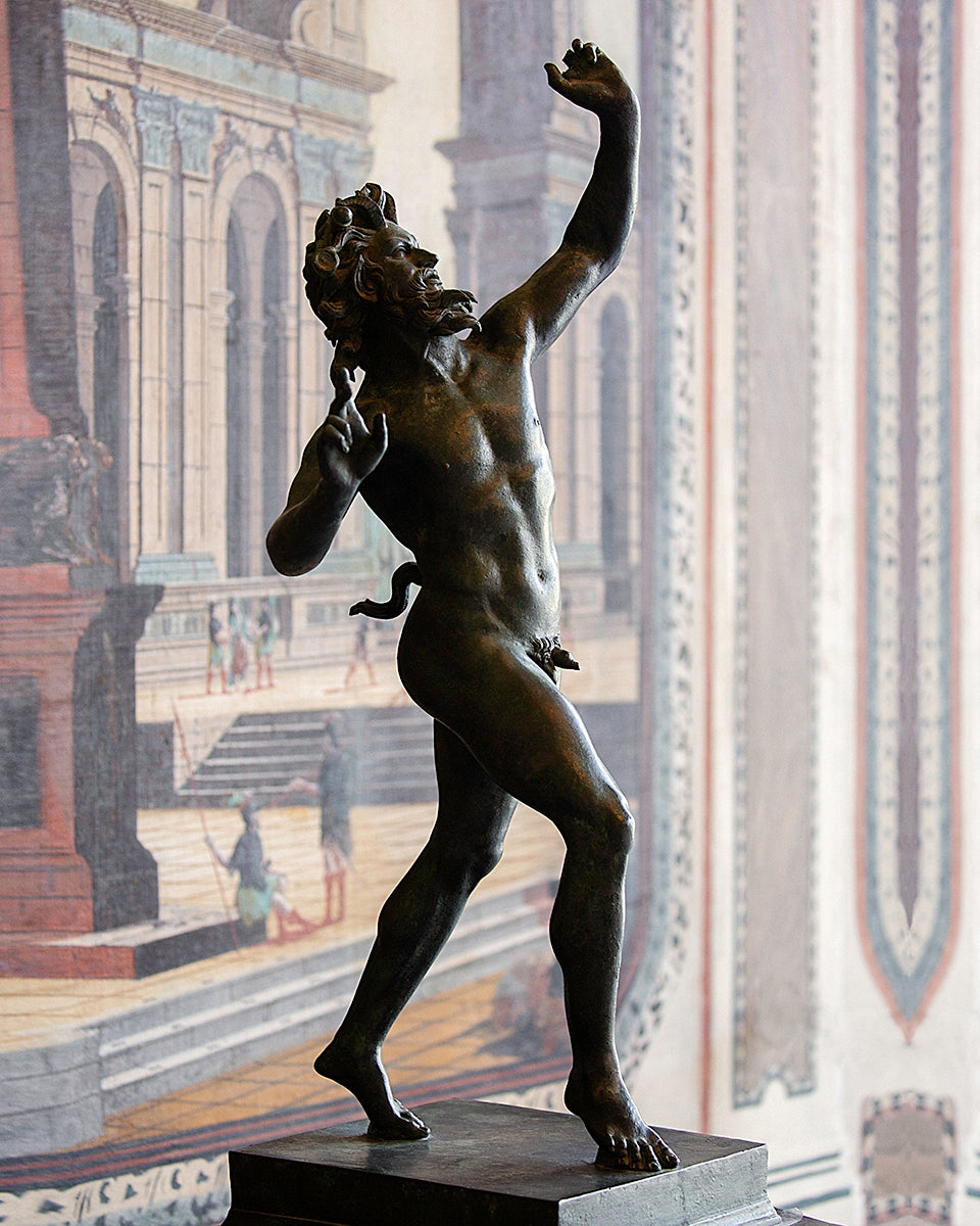 A bronze statue of a nude male figure in a dynamic pose, mounted on a dark pedestal in front of a large classical architectural mural.