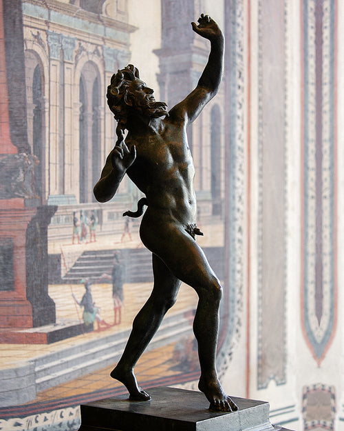 A bronze statue of a nude male figure in a dynamic pose, mounted on a dark pedestal in front of a large classical architectural mural.