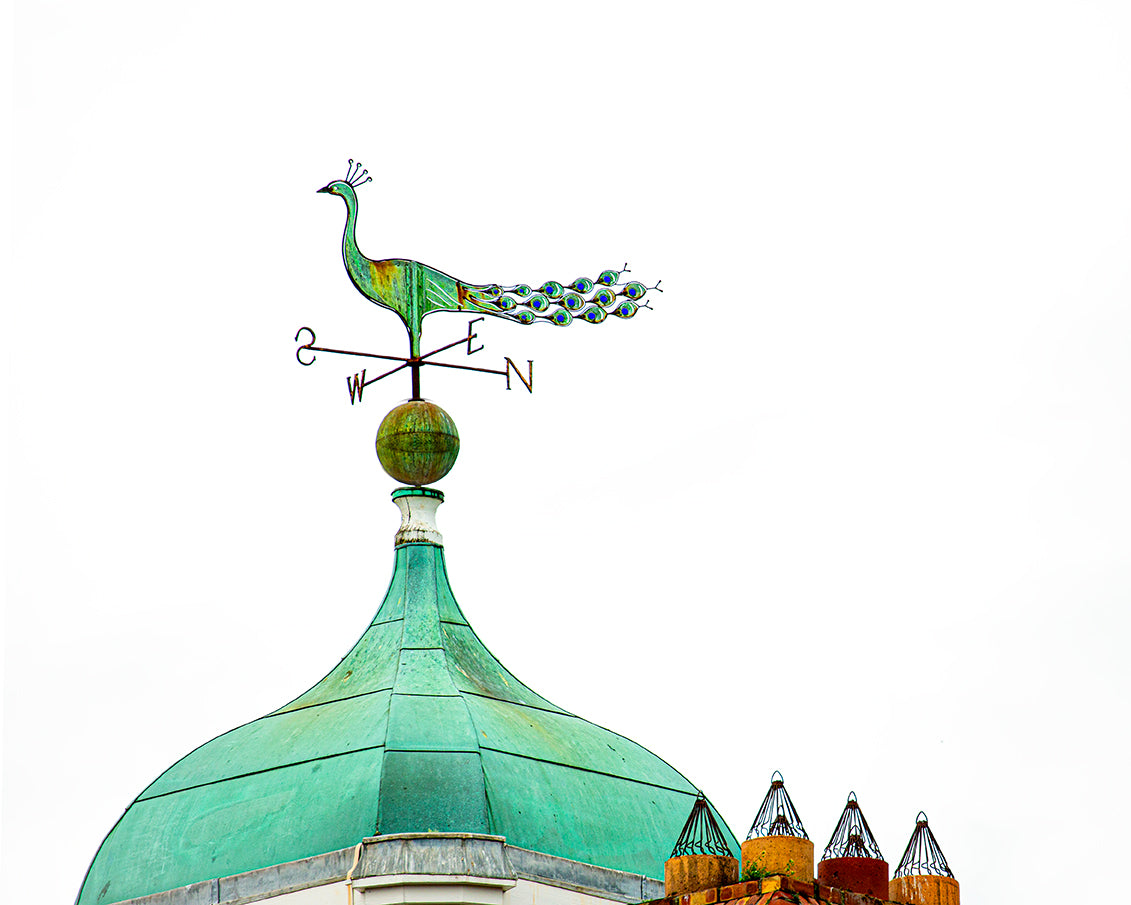 A decorative peacock weathervane mounted on a green copper dome roof against a bright white sky